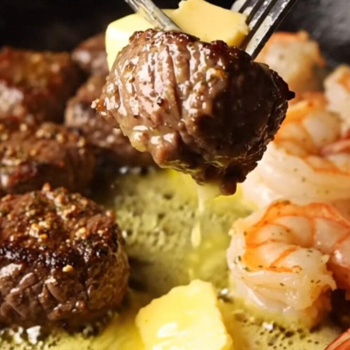 A cast-iron skillet filled with seared steak bites and jumbo shrimp in a golden lemon herb butter sauce.