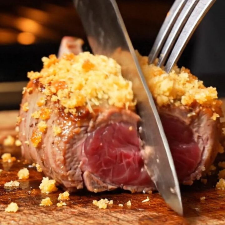 A close-up of golden garlic parmesan crusted lamb loin chops in a cast-iron skillet with ruby-pink centers.