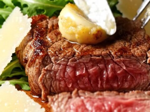 A close-up of sliced medium-rare bavette steak topped with soft garlic cloves and parmesan.