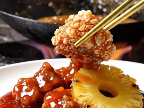 A close-up shot of crispy, golden-brown pork bites tossed in a glossy, vibrant red sweet and sour sauce with pineapple.