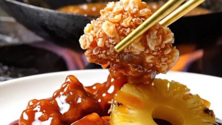 A close-up shot of crispy, golden-brown pork bites tossed in a glossy, vibrant red sweet and sour sauce with pineapple.