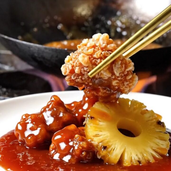 A close-up shot of crispy, golden-brown pork bites tossed in a glossy, vibrant red sweet and sour sauce with pineapple.