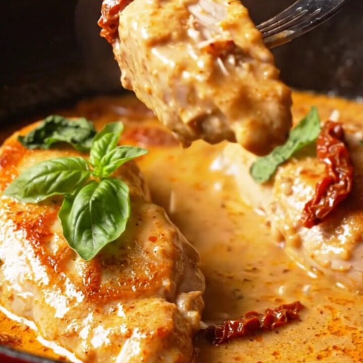 A close-up of golden-seared chicken breast cutlets in a rich, velvety red-pepper and parmesan cream sauce in a cast-iron pan.