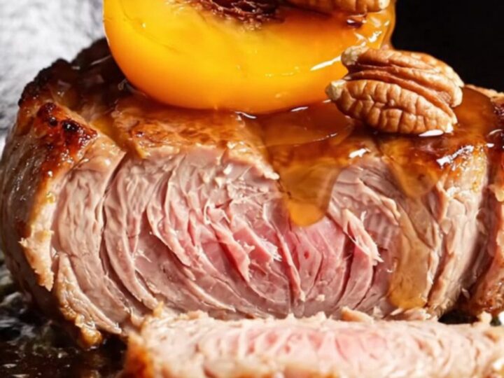 A close-up of thick-cut pork chops topped with caramelized peach wedges and toasted pecans in a cast-iron skillet.