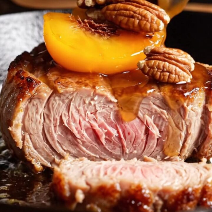 A close-up of thick-cut pork chops topped with caramelized peach wedges and toasted pecans in a cast-iron skillet.