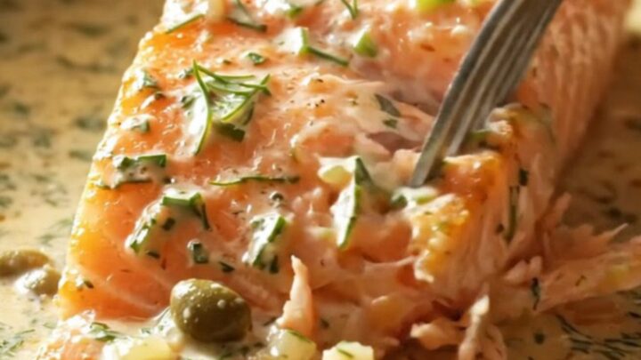 Close-up of a perfectly seared salmon fillet covered in a rich cream sauce with fresh dill and capers.