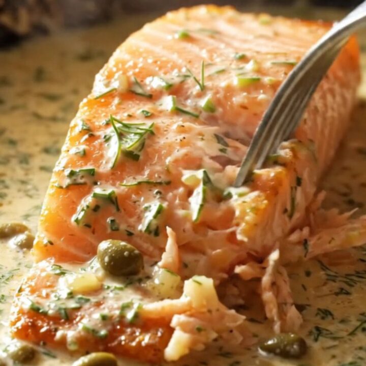 Close-up of a perfectly seared salmon fillet covered in a rich cream sauce with fresh dill and capers.