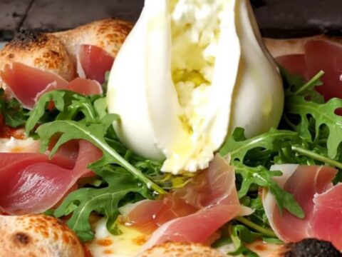 A close-up shot of a wood-fired pizza with a charred, leopard-spotted crust topped with creamy burrata and prosciutto.