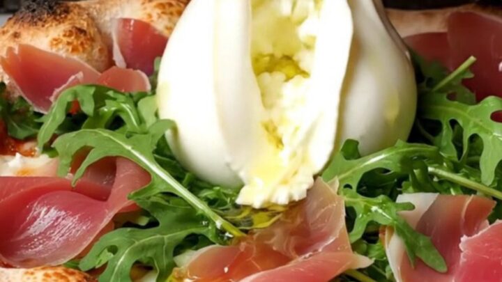 A close-up shot of a wood-fired pizza with a charred, leopard-spotted crust topped with creamy burrata and prosciutto.