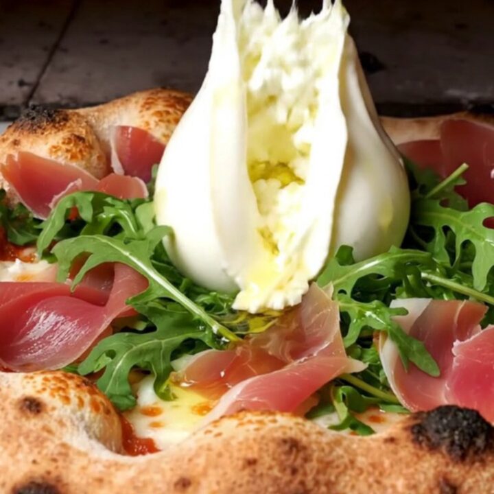 A close-up shot of a wood-fired pizza with a charred, leopard-spotted crust topped with creamy burrata and prosciutto.