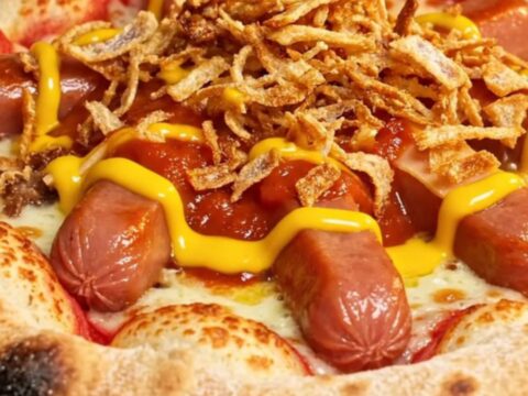 A close-up of a stuffed crust pizza slice being pulled apart, showing melted cheddar and a smoky beef frank inside.
