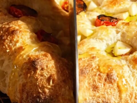 A golden, bubbly focaccia topped with roasted garlic cloves and halved cherry tomatoes on a rustic wooden surface.
