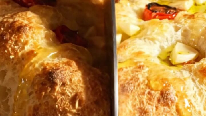 A golden, bubbly focaccia topped with roasted garlic cloves and halved cherry tomatoes on a rustic wooden surface.