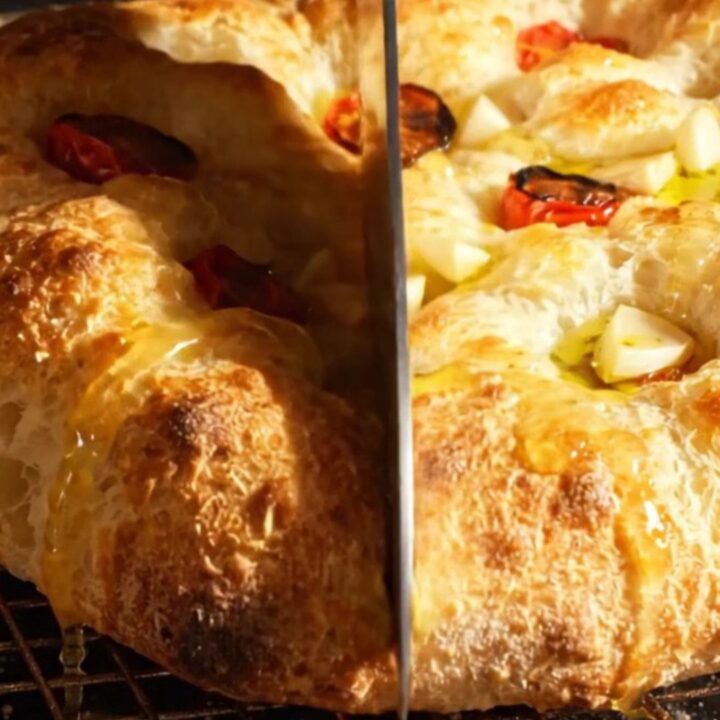 A golden, bubbly focaccia topped with roasted garlic cloves and halved cherry tomatoes on a rustic wooden surface.