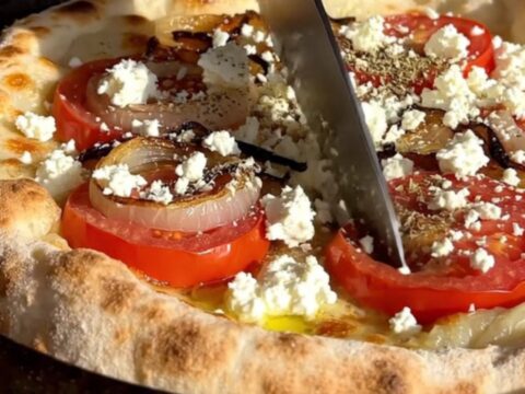A rustic Greek Ladenia flatbread with tomatoes, onions, and feta on a wooden table.