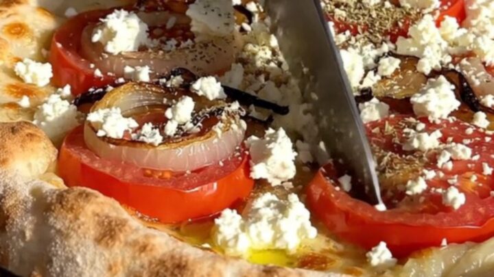 A rustic Greek Ladenia flatbread with tomatoes, onions, and feta on a wooden table.