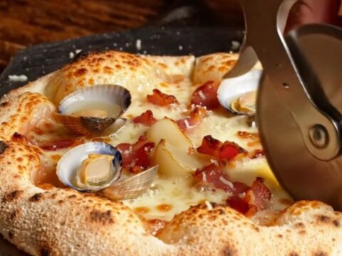 A close-up of a charred New Haven-style white clam pizza topped with bacon and fresh lemon.