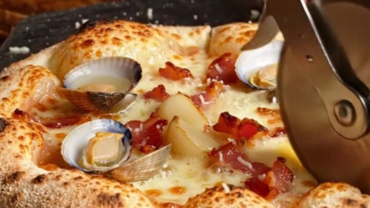 A close-up of a charred New Haven-style white clam pizza topped with bacon and fresh lemon.
