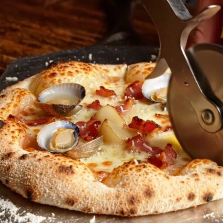 A close-up of a charred New Haven-style white clam pizza topped with bacon and fresh lemon.