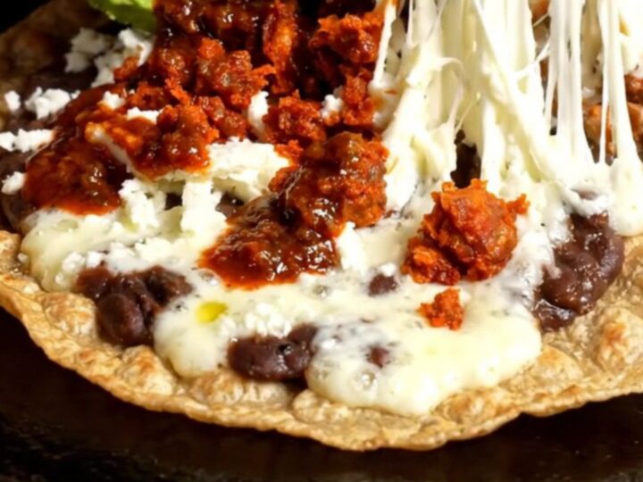 A crispy, golden Oaxacan Tlayuda topped with refried beans, chorizo, melted string cheese, avocado, and salsa roja.