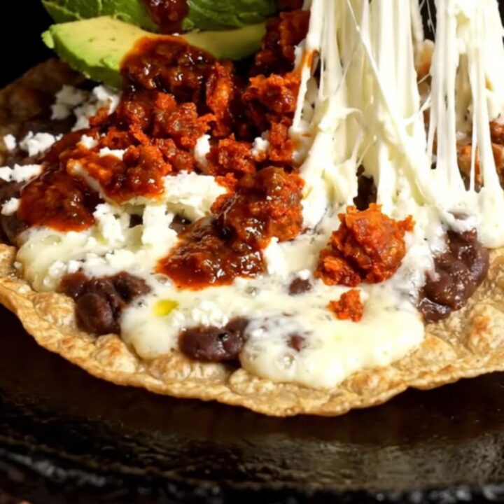 A crispy, golden Oaxacan Tlayuda topped with refried beans, chorizo, melted string cheese, avocado, and salsa roja.