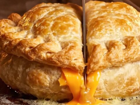 A golden, hand-crimped beef and cheddar pasty cut in half, revealing a steaming