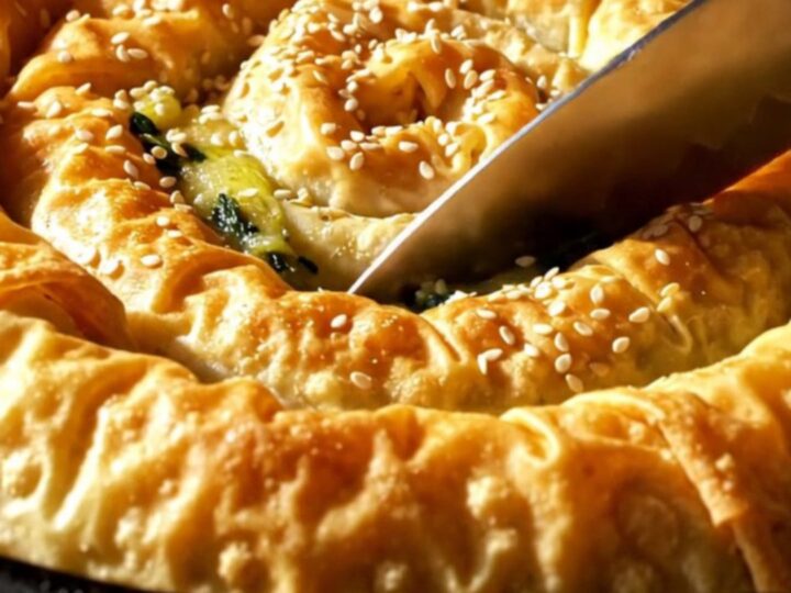 A golden, crispy spanakopita spiral in a cast-iron skillet, topped with toasted sesame seeds.