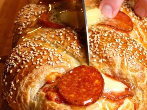 A close-up of a golden, sesame-crusted pepperoni and ricotta stromboli braid with a long cheese pull.