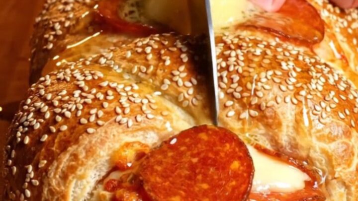 A close-up of a golden, sesame-crusted pepperoni and ricotta stromboli braid with a long cheese pull.