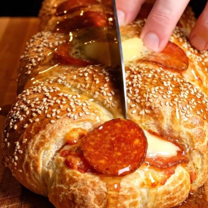 A close-up of a golden, sesame-crusted pepperoni and ricotta stromboli braid with a long cheese pull.
