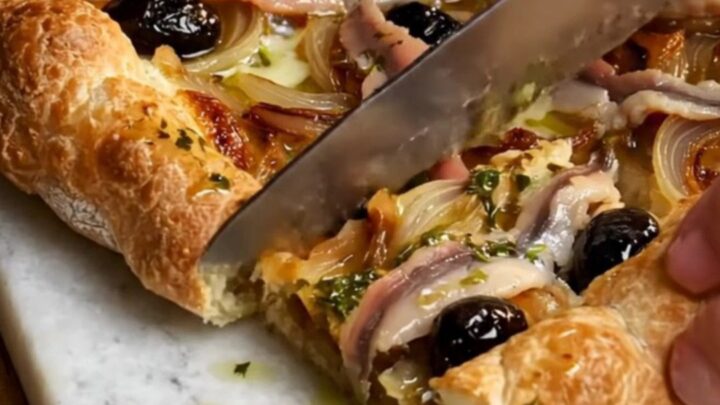 A rustic French Pissaladière tart cut into squares on a wooden board, showing jammy onions and anchovy lattices.