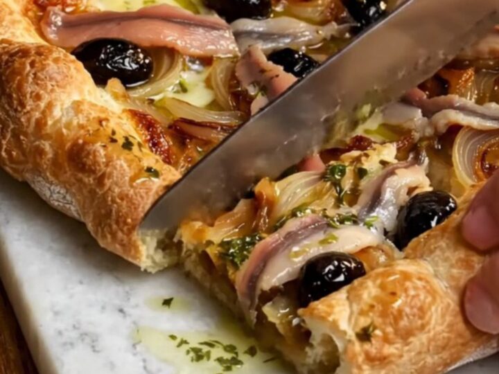 A rustic French Pissaladière tart cut into squares on a wooden board, showing jammy onions and anchovy lattices.