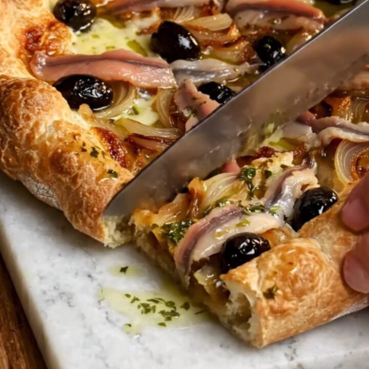 A rustic French Pissaladière tart cut into squares on a wooden board, showing jammy onions and anchovy lattices.