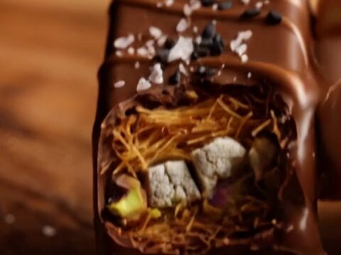 A close-up of a dark chocolate bar broken in half, revealing a crunchy golden kataifi and halva filling.