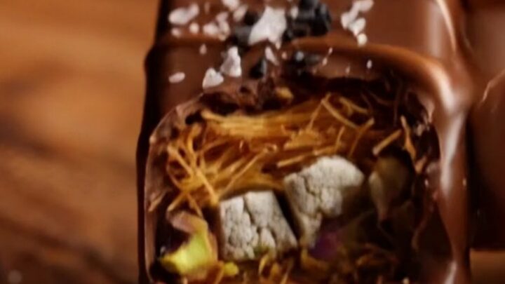 A close-up of a dark chocolate bar broken in half, revealing a crunchy golden kataifi and halva filling.