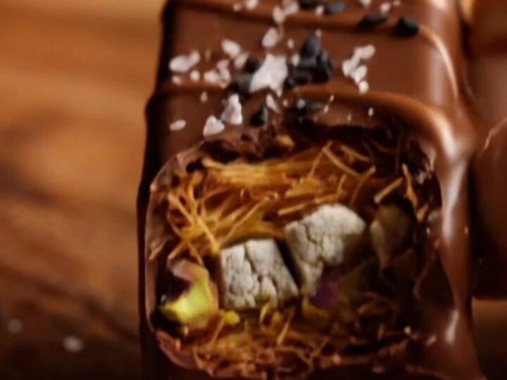 A close-up of a dark chocolate bar broken in half, revealing a crunchy golden kataifi and halva filling.