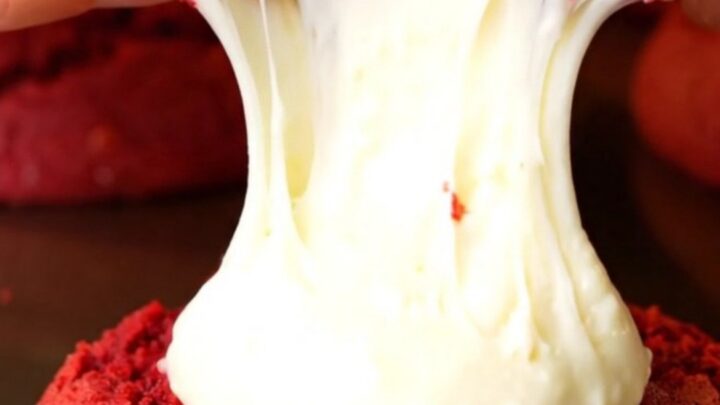 A close-up of a red velvet cookie being pulled apart to show a molten cream cheese center.