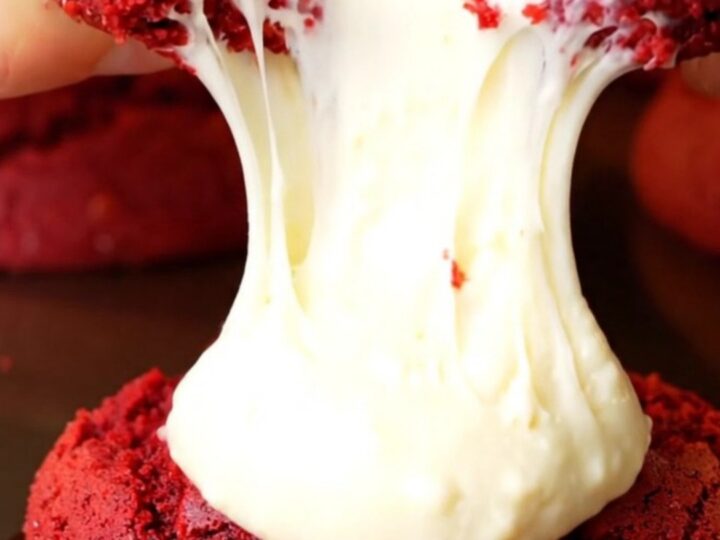 A close-up of a red velvet cookie being pulled apart to show a molten cream cheese center.