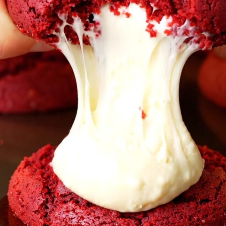 A close-up of a red velvet cookie being pulled apart to show a molten cream cheese center.