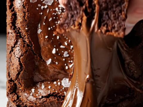 A close-up shot of a square of dark chocolate brownie being lifted with a spatula, revealing a glossy crinkle top.
