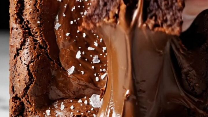 A close-up shot of a square of dark chocolate brownie being lifted with a spatula, revealing a glossy crinkle top.