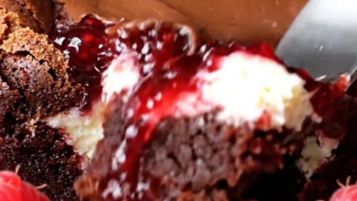 A close-up of a dark fudge brownie with visible swirls of white cheesecake and bright red raspberry jam.