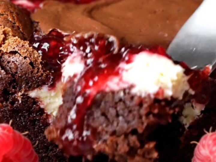 A close-up of a dark fudge brownie with visible swirls of white cheesecake and bright red raspberry jam.