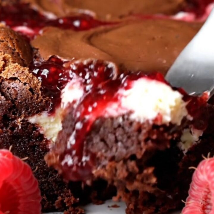 A close-up of a dark fudge brownie with visible swirls of white cheesecake and bright red raspberry jam.