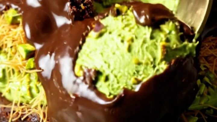A close-up shot showing the layers of a decadent brownie topped with green pistachio kataifi and a dark chocolate shell.