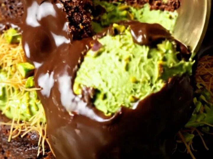 A close-up shot showing the layers of a decadent brownie topped with green pistachio kataifi and a dark chocolate shell.