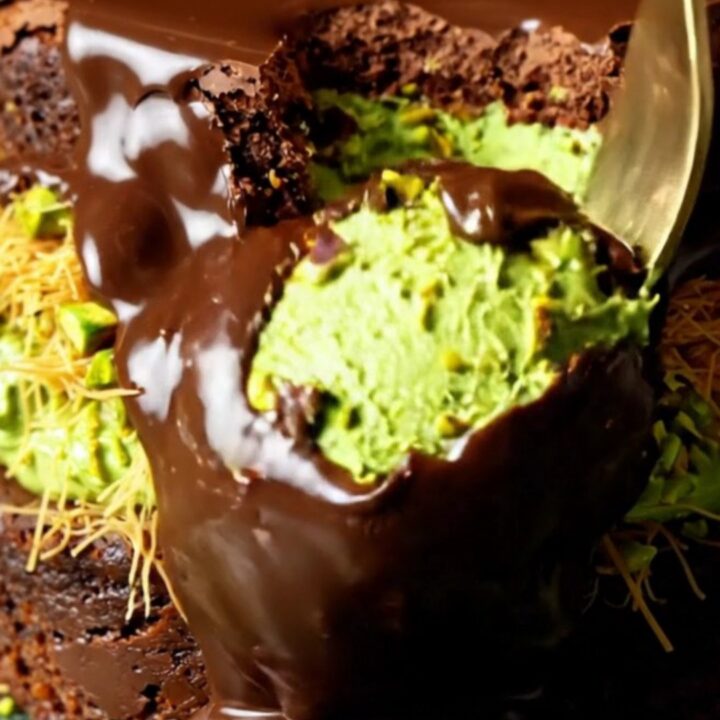 A close-up shot showing the layers of a decadent brownie topped with green pistachio kataifi and a dark chocolate shell.