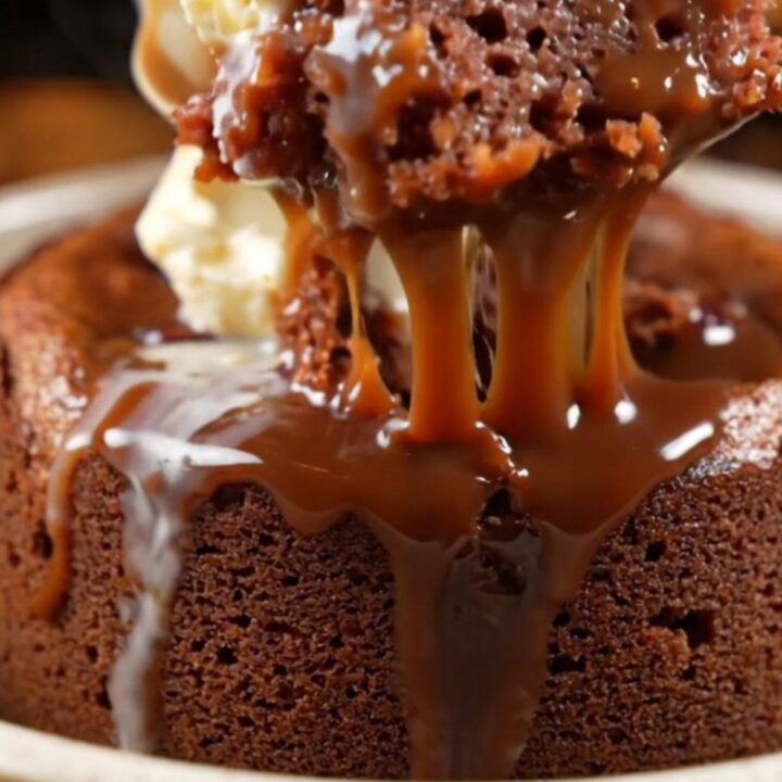 A close-up of dark chocolate toffee sauce being poured over a warm, spongy date and chocolate pudding cake.