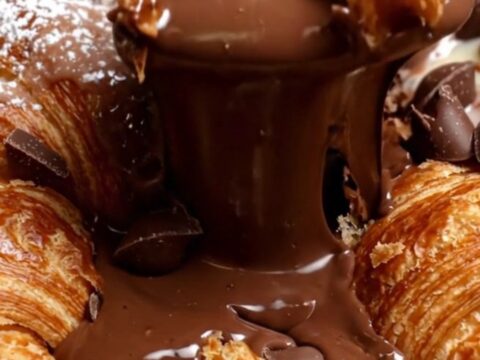 A close-up of a spoon breaking the crispy, golden-brown crust of a croissant chocolate pudding skillet.