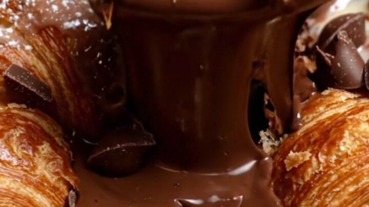A close-up of a spoon breaking the crispy, golden-brown crust of a croissant chocolate pudding skillet.
